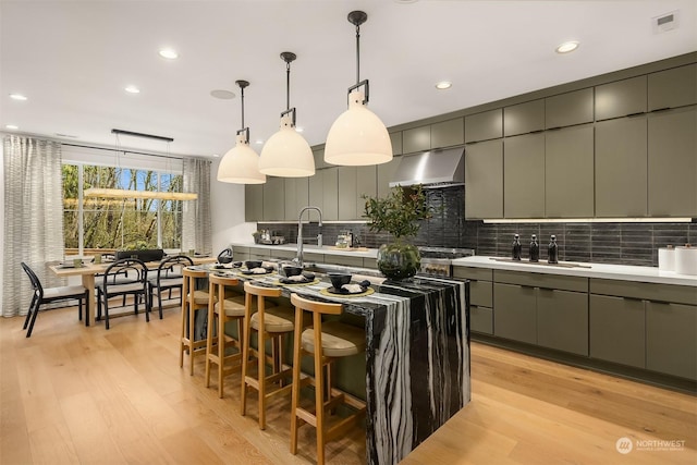 kitchen with ventilation hood, tasteful backsplash, an island with sink, sink, and hanging light fixtures