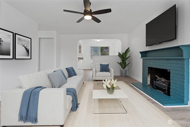 living room with a brick fireplace, a textured ceiling, ceiling fan, and light hardwood / wood-style floors