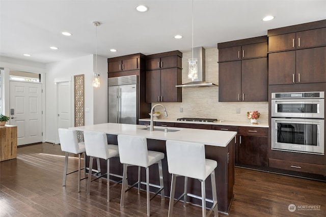 kitchen featuring decorative light fixtures, sink, stainless steel appliances, a center island with sink, and wall chimney exhaust hood