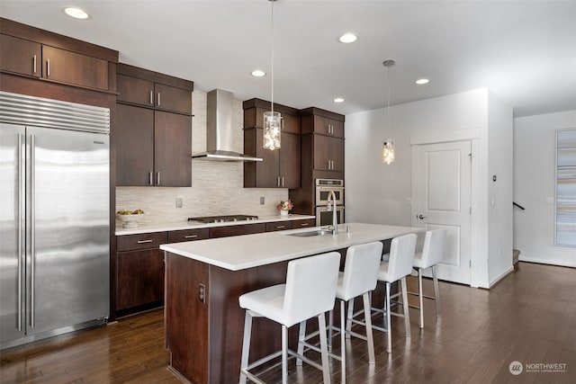 kitchen featuring dark wood-type flooring, a center island with sink, appliances with stainless steel finishes, pendant lighting, and wall chimney range hood