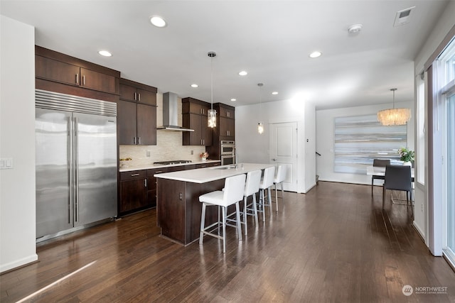 kitchen with hanging light fixtures, stainless steel appliances, dark wood-type flooring, a center island with sink, and wall chimney exhaust hood