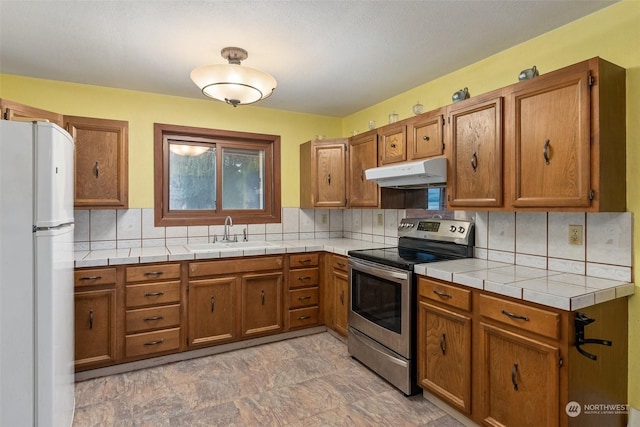 kitchen featuring sink, stainless steel range with electric stovetop, tile countertops, white refrigerator, and backsplash