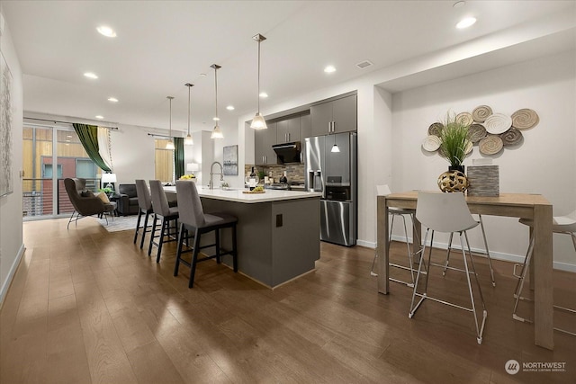 kitchen featuring a breakfast bar area, stainless steel fridge, a kitchen island with sink, hanging light fixtures, and dark hardwood / wood-style flooring