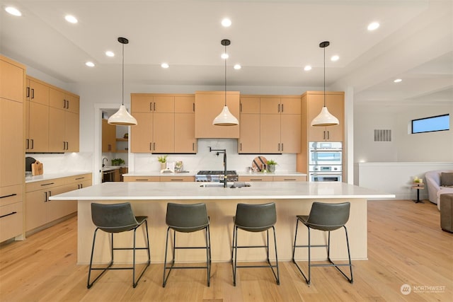 kitchen with an island with sink, light wood-type flooring, light brown cabinets, and double oven