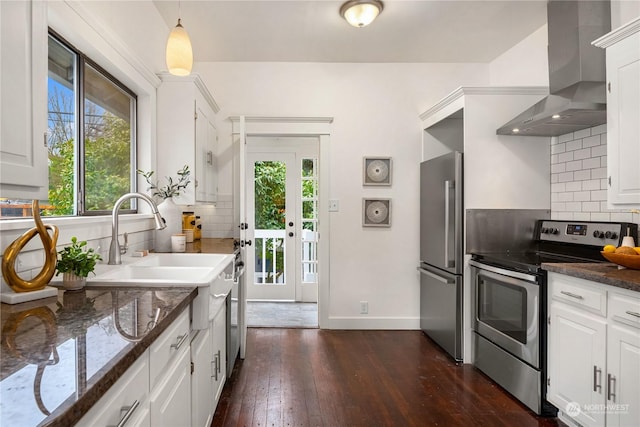 kitchen featuring wall chimney range hood, appliances with stainless steel finishes, hanging light fixtures, white cabinets, and dark stone counters