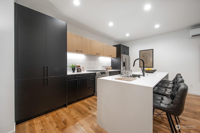 kitchen with appliances with stainless steel finishes, an island with sink, a kitchen breakfast bar, light brown cabinets, and light wood-type flooring