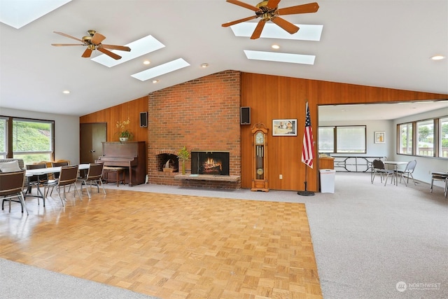 living room with a skylight, high vaulted ceiling, a brick fireplace, wooden walls, and ceiling fan