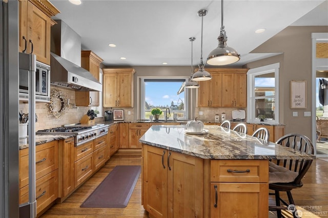 kitchen featuring a breakfast bar, light stone counters, appliances with stainless steel finishes, a kitchen island, and wall chimney range hood