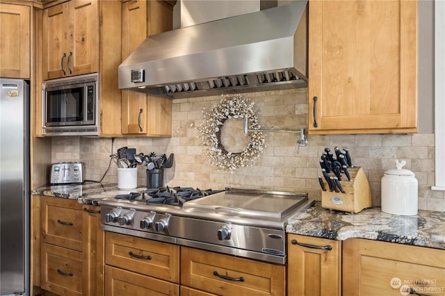 kitchen with dark stone countertops, appliances with stainless steel finishes, wall chimney range hood, and backsplash