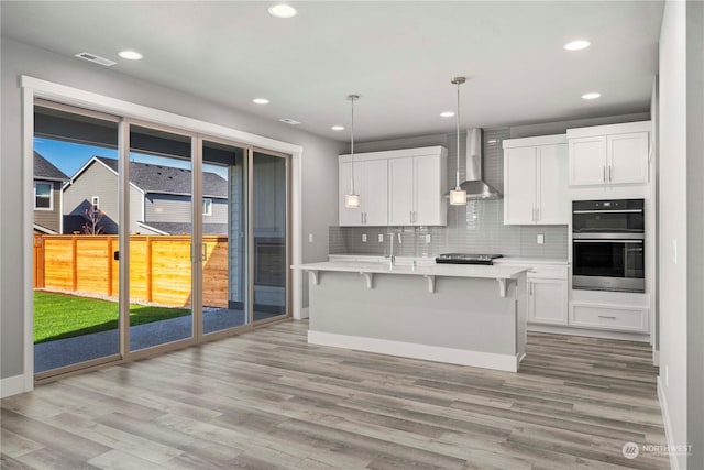 kitchen featuring double oven, decorative light fixtures, an island with sink, white cabinets, and wall chimney exhaust hood