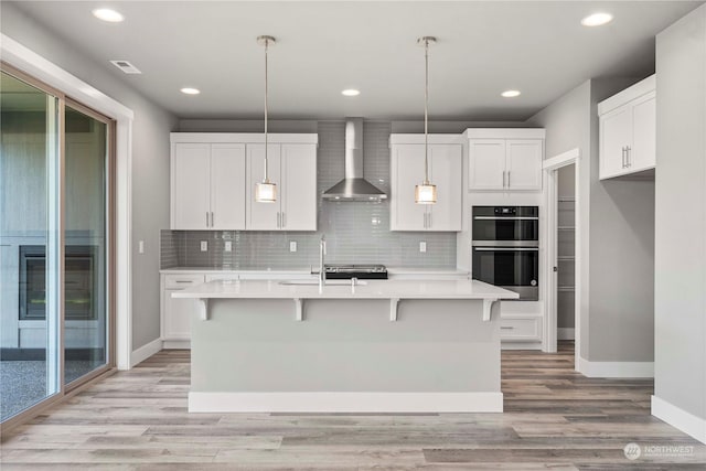 kitchen featuring hanging light fixtures, white cabinetry, a center island with sink, and wall chimney range hood