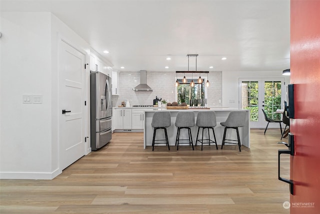 kitchen featuring stainless steel fridge, a breakfast bar area, hanging light fixtures, white cabinets, and wall chimney exhaust hood