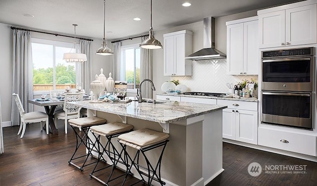 kitchen featuring pendant lighting, wall chimney exhaust hood, white cabinets, a center island with sink, and stainless steel double oven