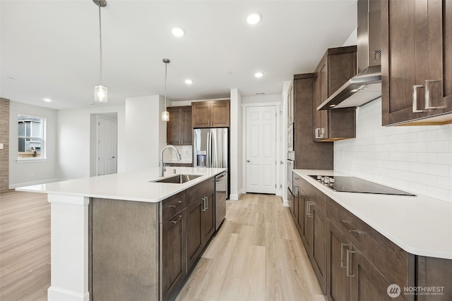 kitchen with pendant lighting, an island with sink, sink, stainless steel appliances, and wall chimney exhaust hood