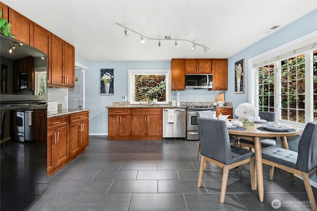 kitchen with stainless steel appliances, brown cabinetry, track lighting, and light stone countertops