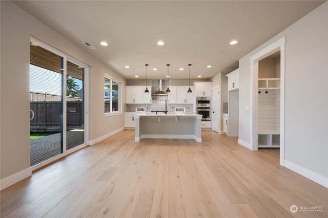 kitchen featuring wall chimney range hood, a kitchen island with sink, stainless steel appliances, white cabinets, and decorative light fixtures