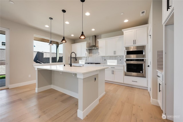 kitchen featuring wall chimney range hood, sink, white cabinetry, hanging light fixtures, and an island with sink