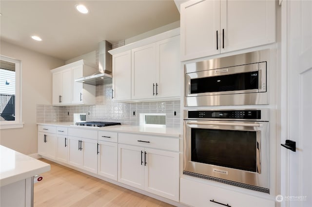 kitchen featuring wall chimney exhaust hood, appliances with stainless steel finishes, white cabinets, light hardwood / wood-style floors, and backsplash