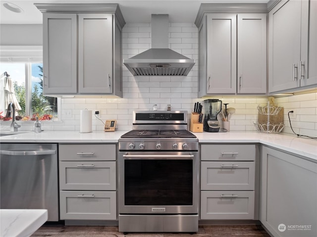 kitchen with gray cabinets, stainless steel appliances, light stone counters, decorative backsplash, and wall chimney exhaust hood