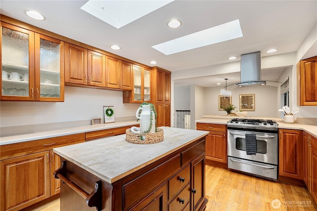 kitchen featuring light wood-style flooring, a skylight, stainless steel gas range oven, brown cabinets, and island range hood