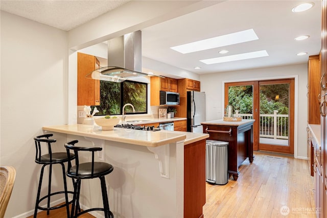 kitchen featuring a peninsula, brown cabinets, a skylight, island range hood, and appliances with stainless steel finishes