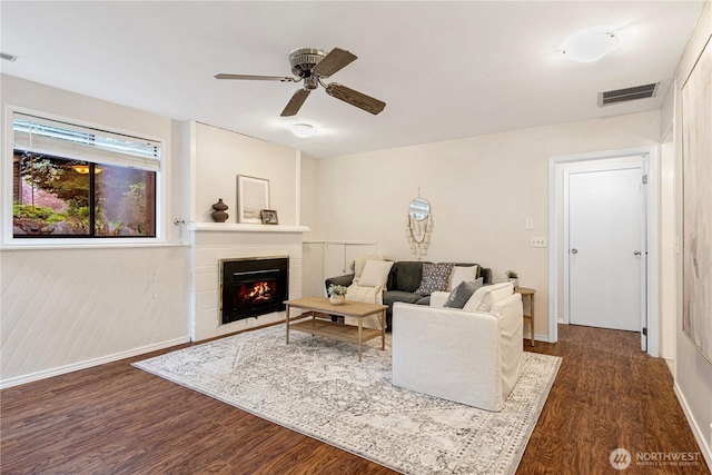 living area featuring visible vents, baseboards, a warm lit fireplace, ceiling fan, and dark wood-type flooring