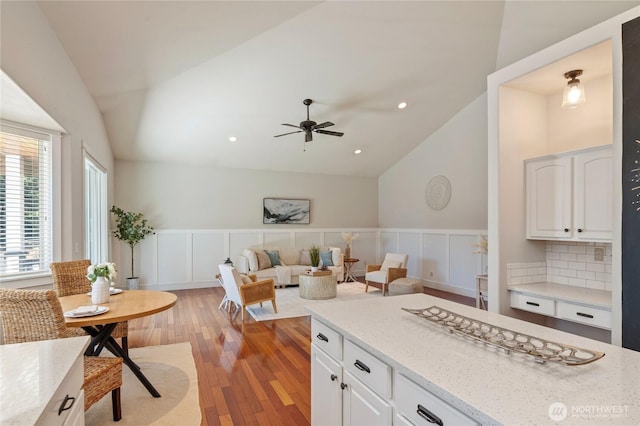 kitchen with a wainscoted wall, white cabinetry, vaulted ceiling, light wood-type flooring, and light stone countertops