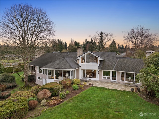 back house at dusk featuring a patio, a sunroom, and a lawn