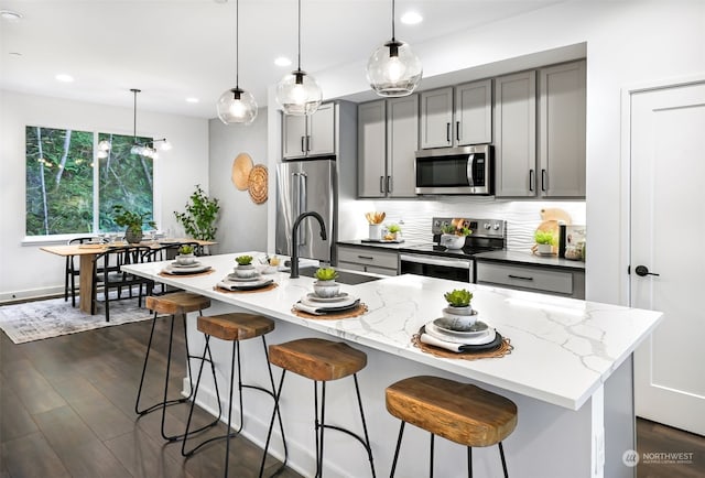 kitchen featuring gray cabinetry, hanging light fixtures, stainless steel appliances, light stone counters, and a center island with sink