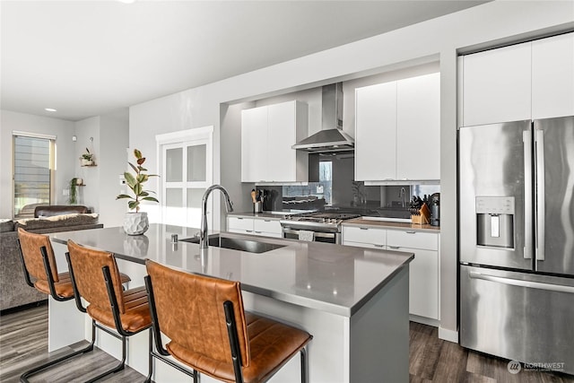 kitchen with sink, a breakfast bar area, a center island with sink, appliances with stainless steel finishes, and wall chimney range hood