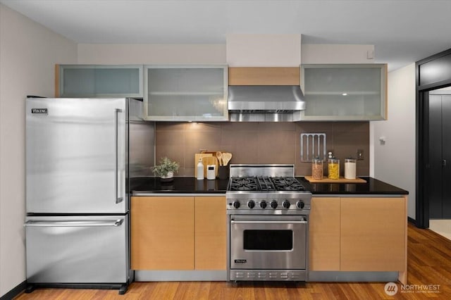 kitchen featuring high end appliances, dark countertops, decorative backsplash, wood finished floors, and wall chimney exhaust hood