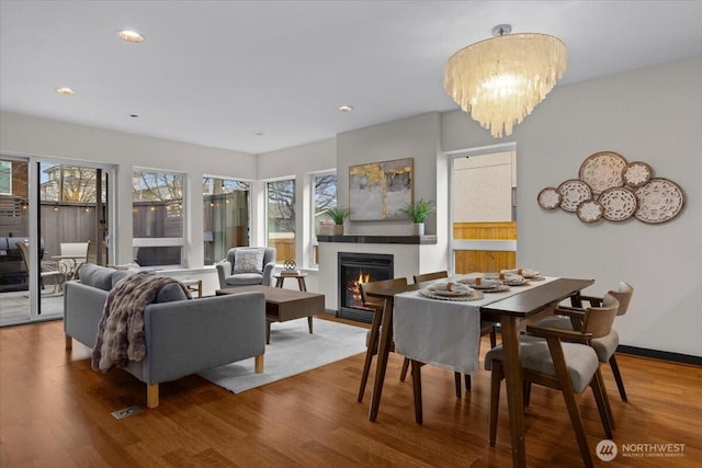 dining room featuring a warm lit fireplace, baseboards, wood finished floors, a notable chandelier, and recessed lighting