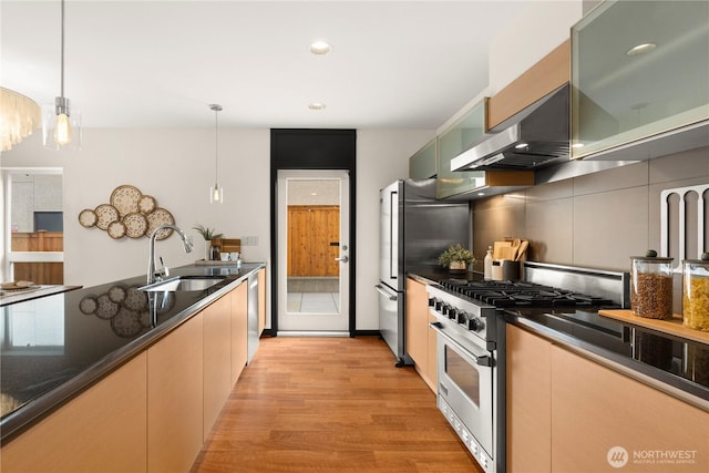 kitchen featuring light wood finished floors, appliances with stainless steel finishes, decorative light fixtures, wall chimney range hood, and a sink