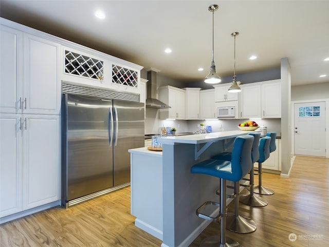 kitchen featuring pendant lighting, wall chimney range hood, light hardwood / wood-style flooring, white cabinetry, and stainless steel built in refrigerator