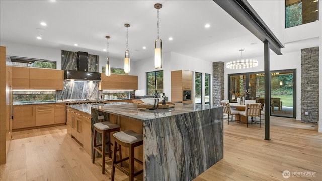 kitchen featuring stone counters, decorative light fixtures, wall chimney range hood, light hardwood / wood-style floors, and a spacious island