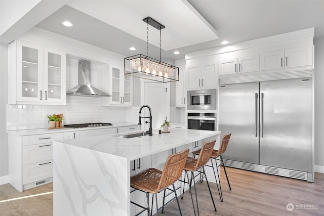 kitchen featuring decorative light fixtures, an island with sink, white cabinets, built in appliances, and wall chimney range hood