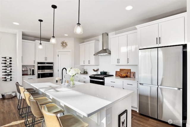 kitchen with pendant lighting, white cabinets, a kitchen island with sink, stainless steel appliances, and wall chimney range hood