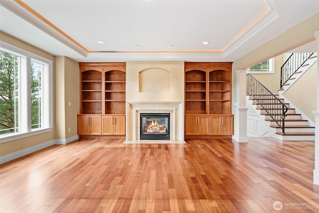 unfurnished living room featuring light wood finished floors, built in features, a tile fireplace, stairs, and a tray ceiling