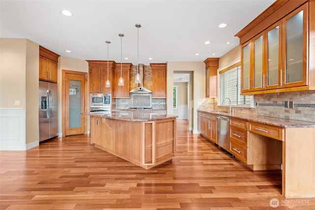 kitchen with a kitchen island, appliances with stainless steel finishes, glass insert cabinets, decorative light fixtures, and wall chimney range hood