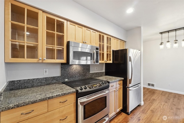 kitchen with appliances with stainless steel finishes, light brown cabinetry, decorative light fixtures, dark stone counters, and light hardwood / wood-style floors