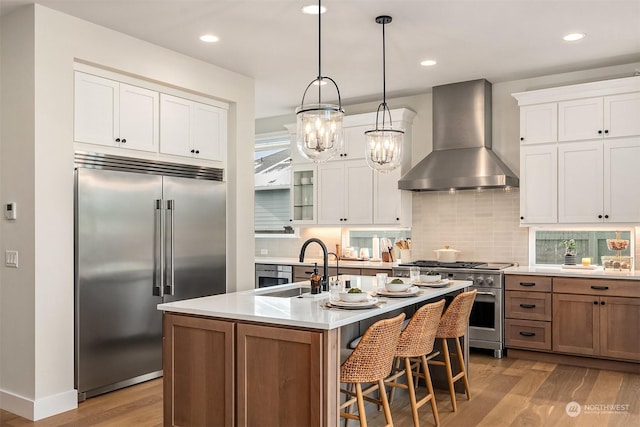 kitchen featuring pendant lighting, wall chimney range hood, white cabinetry, a kitchen island with sink, and high end appliances