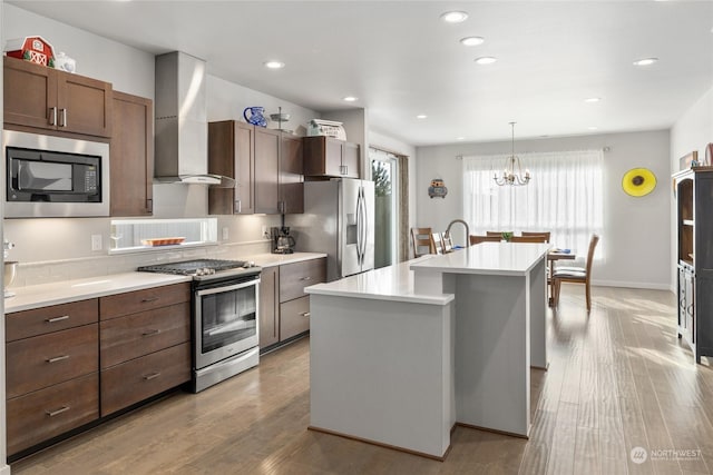 kitchen featuring wall chimney exhaust hood, light hardwood / wood-style flooring, pendant lighting, stainless steel appliances, and a kitchen island with sink