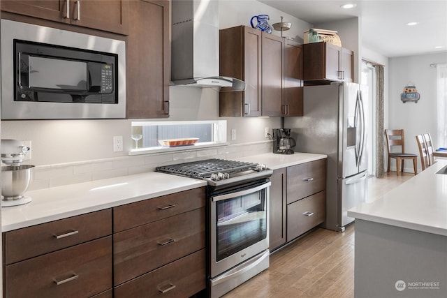 kitchen featuring stainless steel appliances, dark brown cabinets, wall chimney range hood, and light hardwood / wood-style flooring