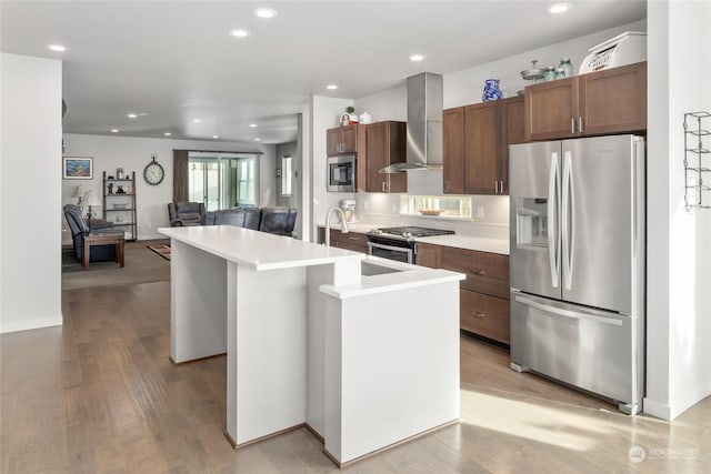 kitchen with sink, stainless steel appliances, an island with sink, wall chimney exhaust hood, and light wood-type flooring