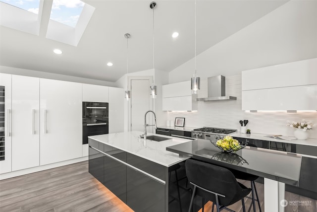 kitchen with white cabinetry, sink, hanging light fixtures, a kitchen island with sink, and wall chimney range hood