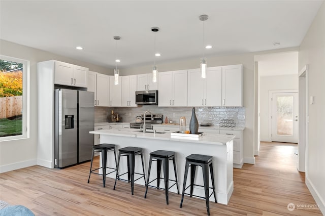 kitchen with white cabinetry, hanging light fixtures, appliances with stainless steel finishes, a kitchen island with sink, and light hardwood / wood-style floors