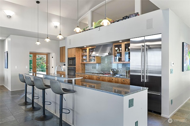 kitchen with dark stone counters, black appliances, wall chimney exhaust hood, hanging light fixtures, and tasteful backsplash