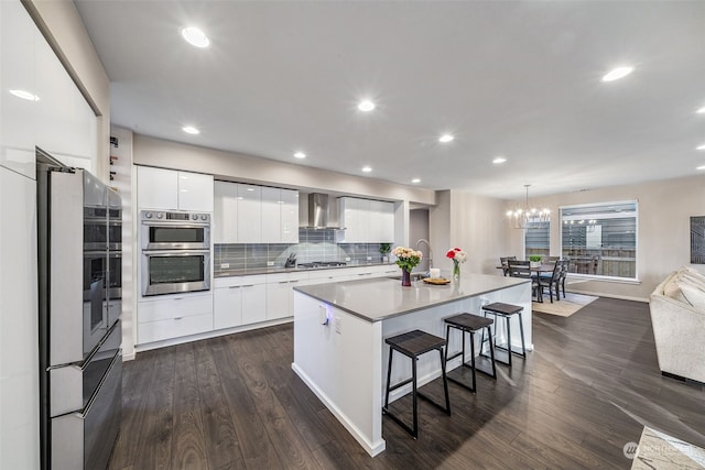 kitchen featuring decorative light fixtures, an island with sink, white cabinets, stainless steel appliances, and wall chimney exhaust hood