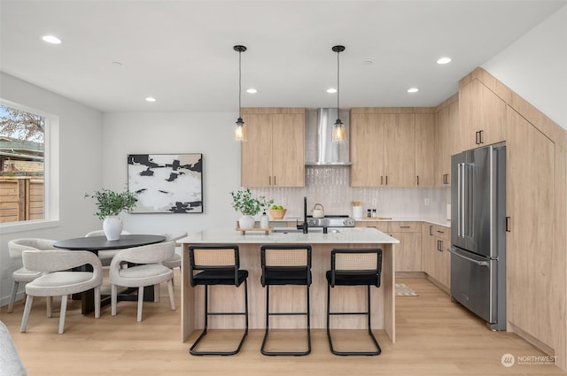 kitchen featuring pendant lighting, an island with sink, high end refrigerator, wall chimney exhaust hood, and light brown cabinets