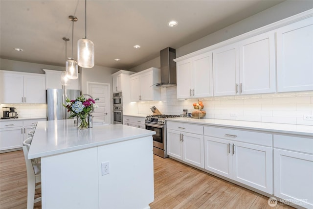 kitchen featuring decorative light fixtures, wall chimney range hood, light hardwood / wood-style floors, stainless steel appliances, and white cabinets